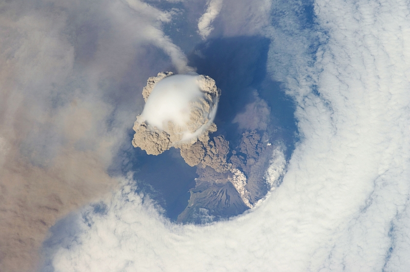 6. Sarychev Peak Volcano Erupts -- Clearing,  Possibly From Its Shock Wave, A Circle In the Cloud Deck Above -- With A Plume of Brown Ash and Steam, June 12, 2009 at 22:16:14 GMT, Matua Island, Kuril Islands, Rossiyskaya Federatsiya -- Russian Federation, As Seen From the International Space Station (Expedition 20) Photo Credit: NASA, International Space Station (Expedition Twenty); ISS020-E-9048, Sarychev Peak, Invisible shock wave, Volcanic plume, Matua Island, Kuril (or Kurile) Islands; Image Science and Analysis Laboratory, NASA-Johnson Space Center. 'Astronaut Photography of Earth - Display Record.' <http://eol.jsc.nasa.gov/scripts/sseop/photo.pl?mission=ISS020&roll=E&frame=9048>; National Aeronautics and Space Administration (NASA, http://www.nasa.gov), Government of the United States of America (USA).