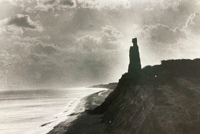 Black and white photo of a rugged coastline with a distinctive rock formation resembling a tower, under a dramatic sky with visible clouds, next to a calm sea.