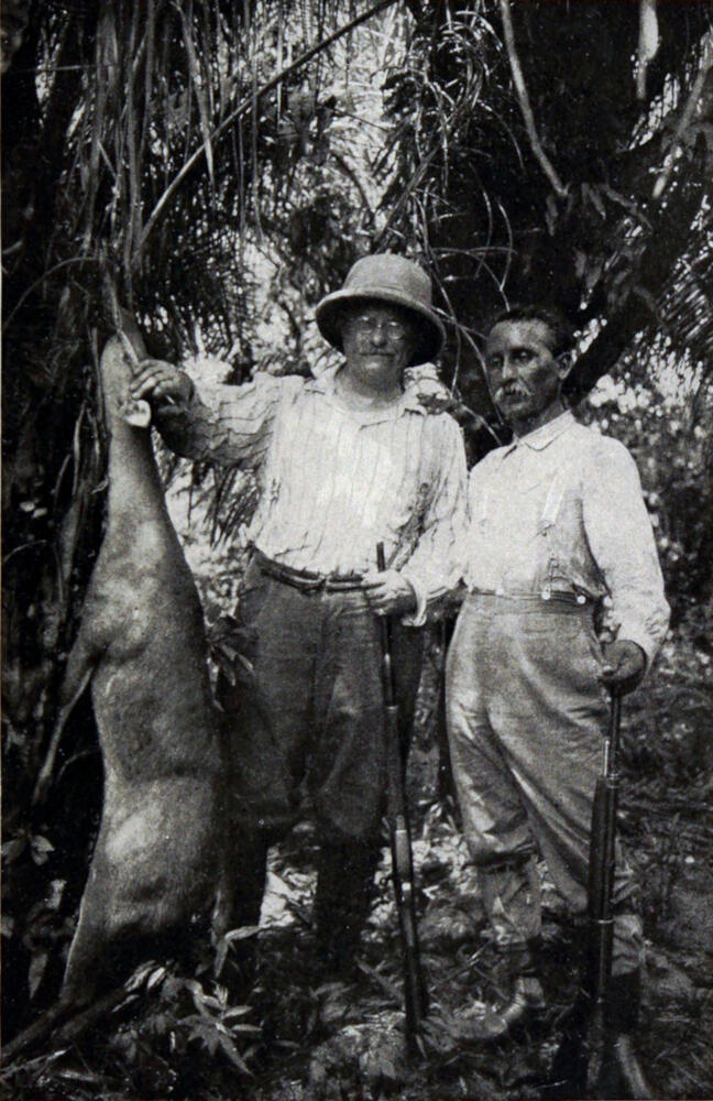A photograph of two men holding rifles and standing next to the body of a deer hanging in a tree. The man on the left is resting his hand on the deer’s head.