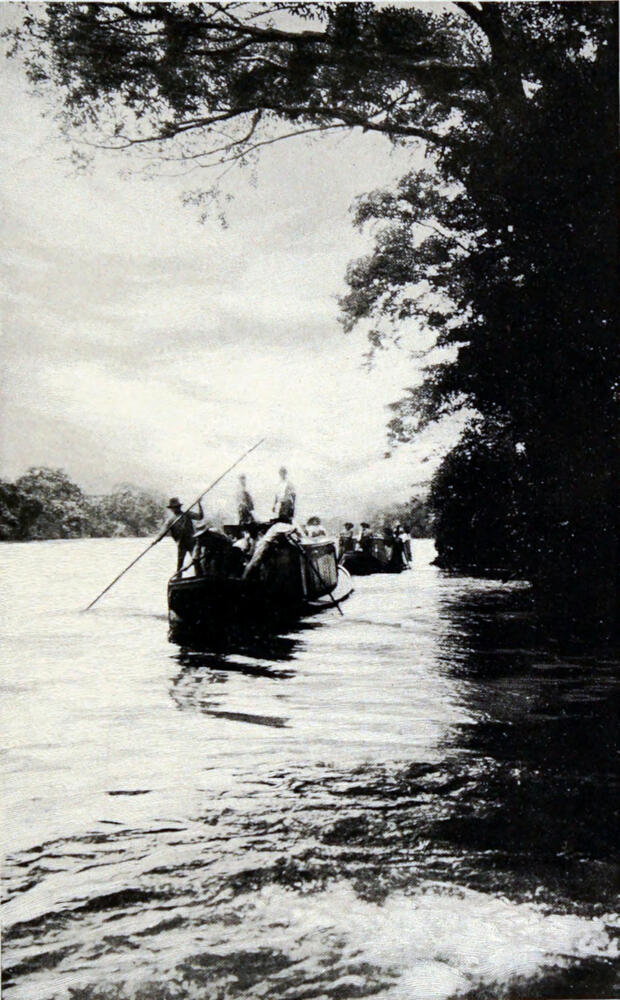 A photograph of a river, with two boats moving away from the camera. Each boat is full of men, and there is a man on the last boat standing up and holding the end a long pole. The other end of the pole is in the water.