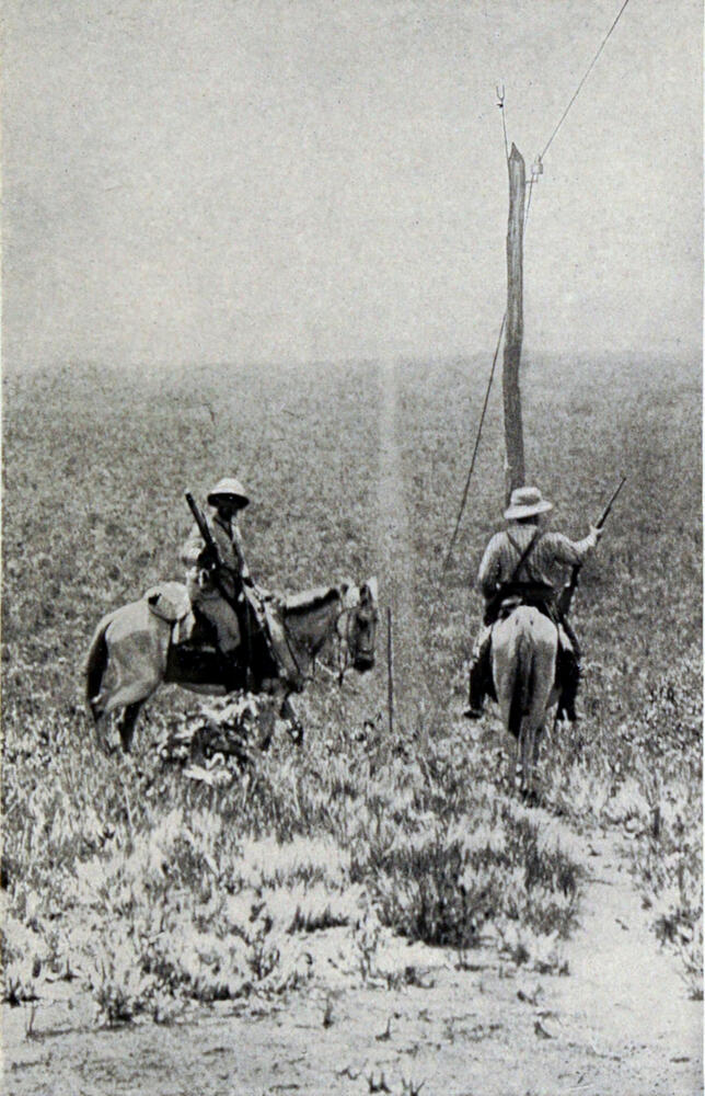 A photograph of two men on horseback carrying rifles. They are standing in the middle of a vast expanse of brushy, sandy ground.