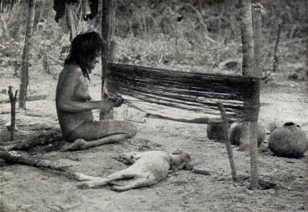 A photograph of a woman kneeling in front of two large upright posts. Long lengths of woven grass are running between the posts as the woman weaves a hammock from them. A dog lies on the ground beside her.