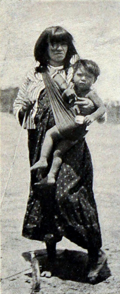 A photograph of a woman carrying a child in a sling draped from her right shoulder across her front.
