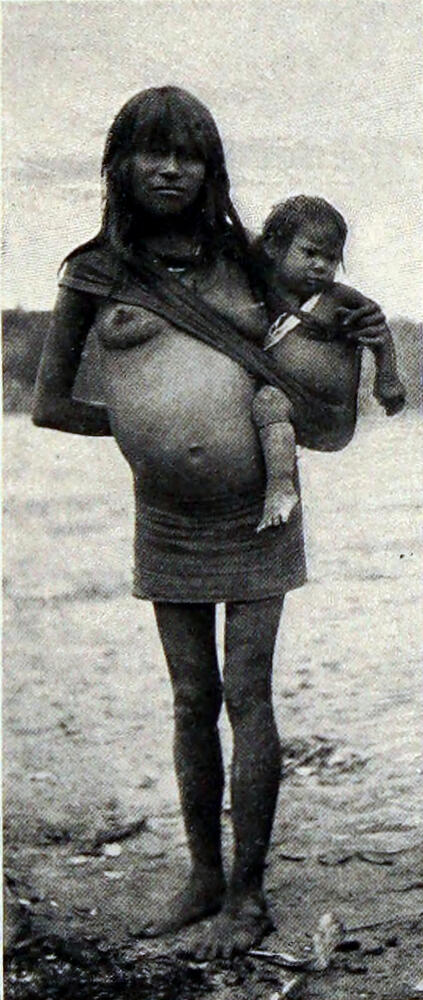 A photograph of a woman carrying a child in a sling draped from her right shoulder, holding the child against her left side.