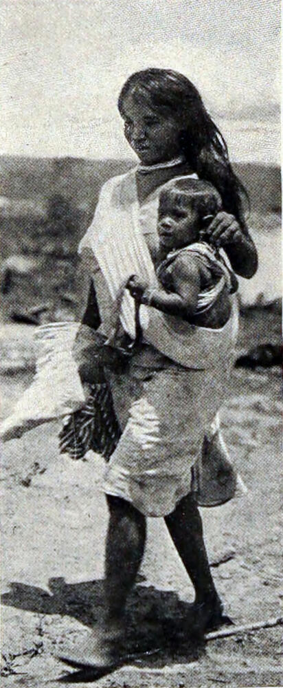 A photograph of a woman carrying a child in a wide, white sling draped from her right shoulder, holding the child against her left side.