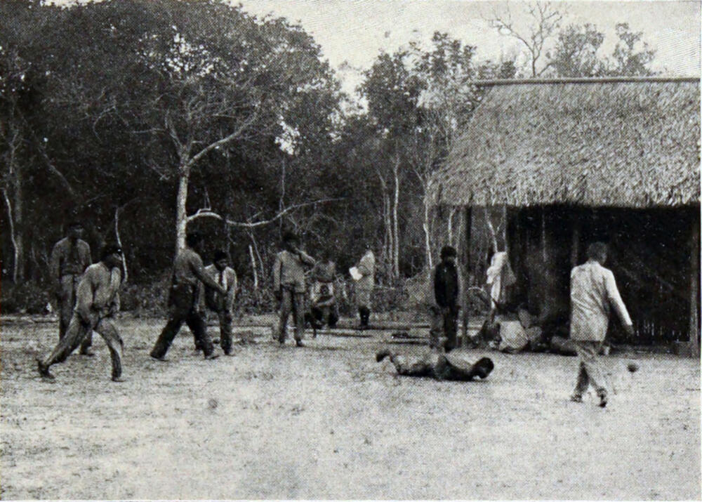 A photograph of twelve men playing a game in front of a building with a thatched roof. One of the men lies on the ground.