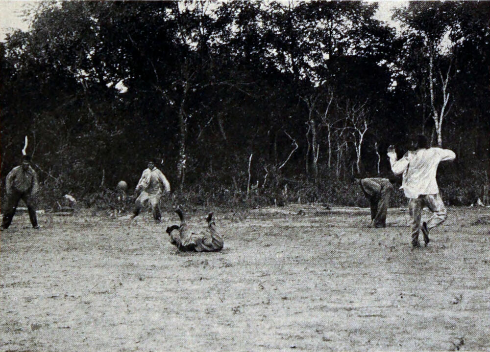 A photograph of five men kicking a ball in a field with numerous trees behind them.