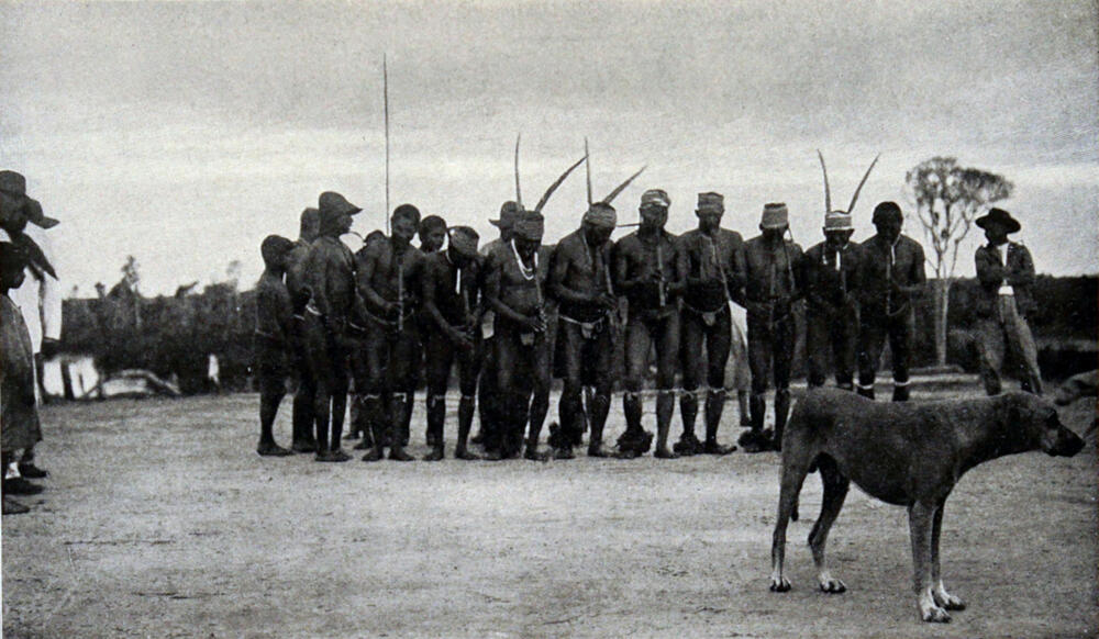 A photograph of numerous men, wearing loincloths, dancing in a circle. A dog stands in front of them.