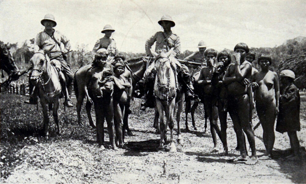A photograph of four men on horseback, with numerous native men, women and children standing among them.