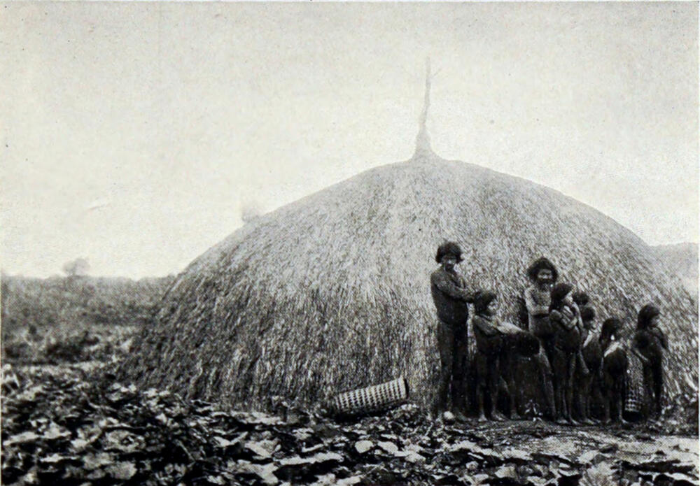 A photograph of a fnative family of nine people standing in front of a dome-shaped hut made of woven grass.