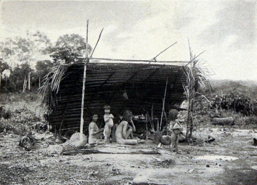 A photograph of a native family sitting under an open-sided shelter made of branches and woven grass.