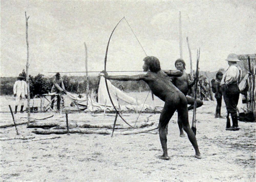 A photograph of a native man standing on sandy ground, holding a bow and very long arrow horizontally. Another native man, holding long spears, stands behind him. A white man, a native woman with a woven basket strapped to her back, and other men are standing nearby.
