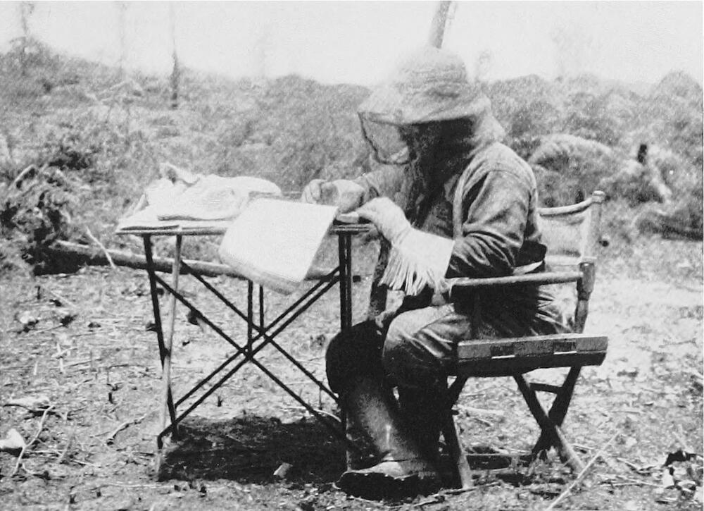 A photograph of a man sitting in a folding chair at a folding table. He is wearing gloves with a fringe down the arm, plas a helmet with a veil.