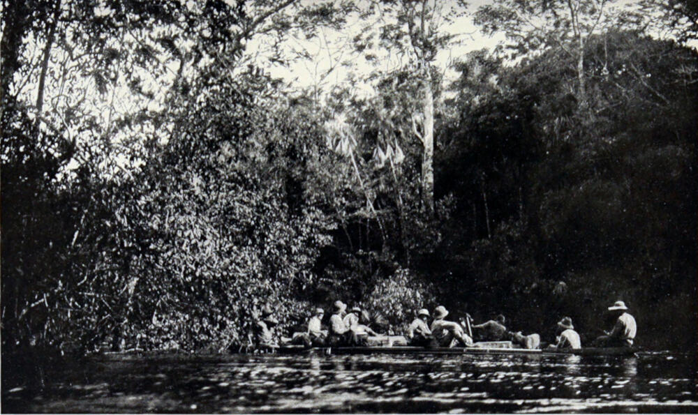 A photograph of a river, with two canoes broadside to the camera containing eight men and one dog.