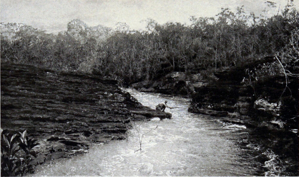 A photograph of a river with rocky banks on either side. A man crouches on the left riverbank and holds a rifle outstretched, almost touching the opposite bank.