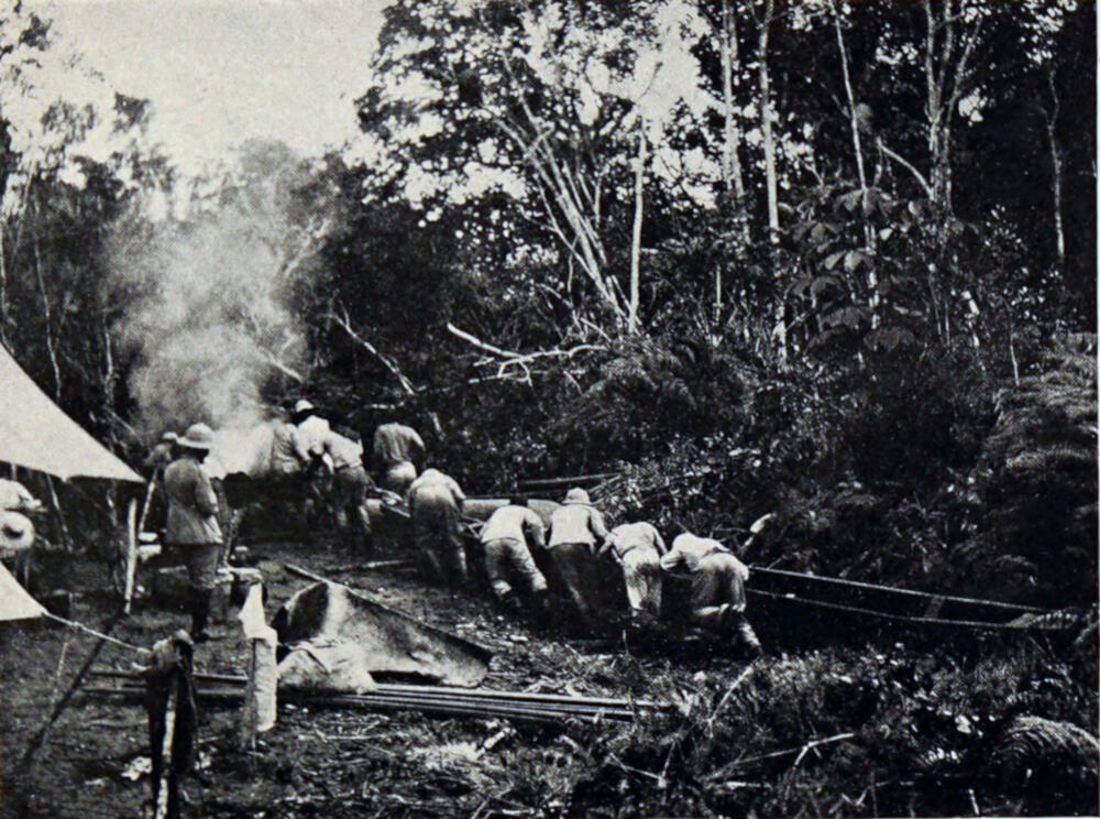A photograph of several men pulling two canoes through a rough jungle area. There is a tent on the left side of the photograph, with smoke rising from a fire near the tent.