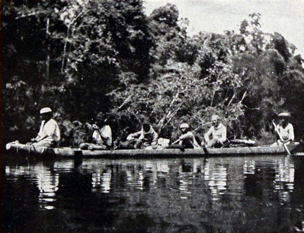A photograph of six men in a canoe. The canoe is broadside to the camera.