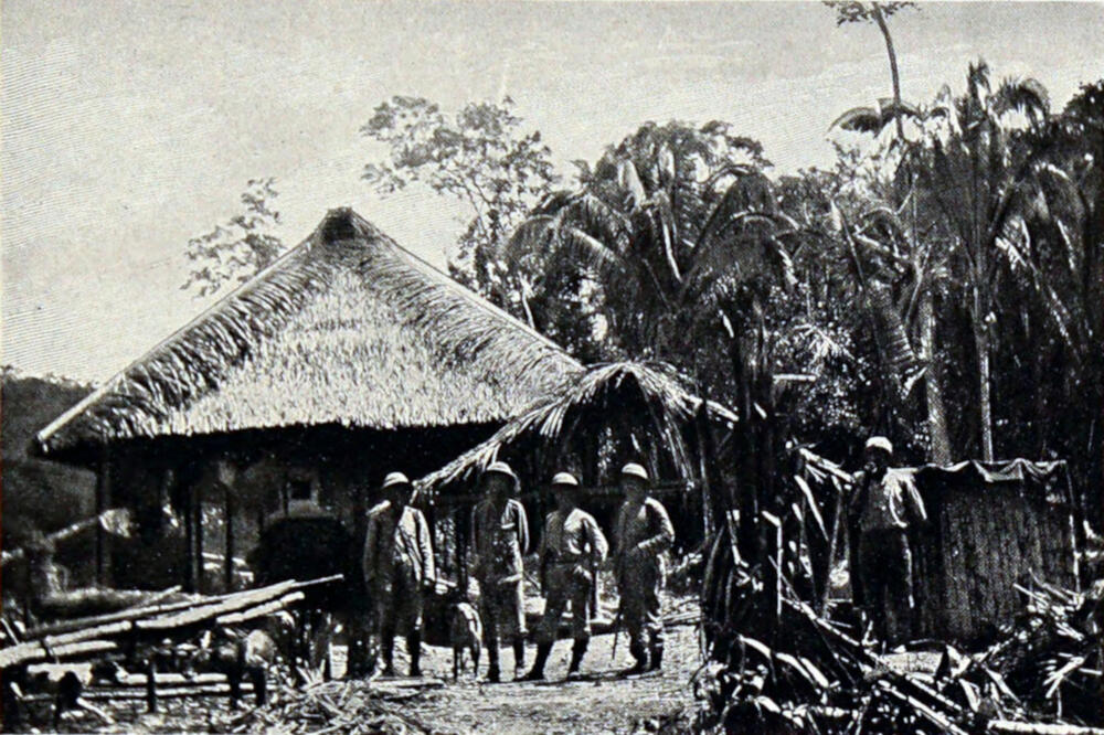 A photograph of five men and one dog standing in front of a hut with a thatched roof.