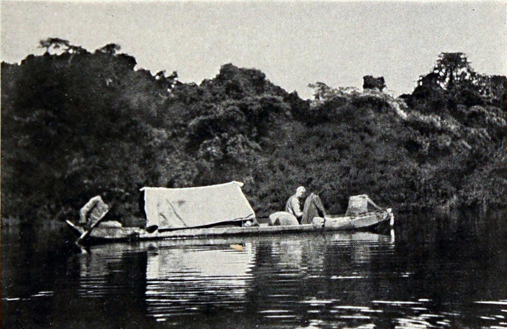 A photograph of three men in a canoe on the water, broadside to the camera, with a tarp on top about halfway to the rear of the canoe.