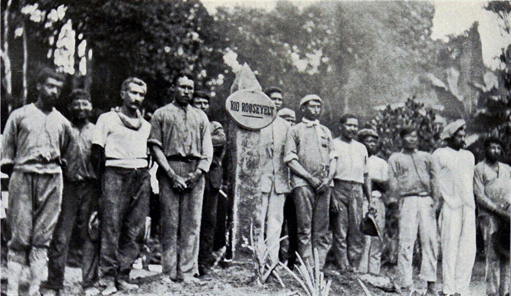 A photograph of thirteen men standing around a pillar that bears a sign that says “Rio Roosevelt.”