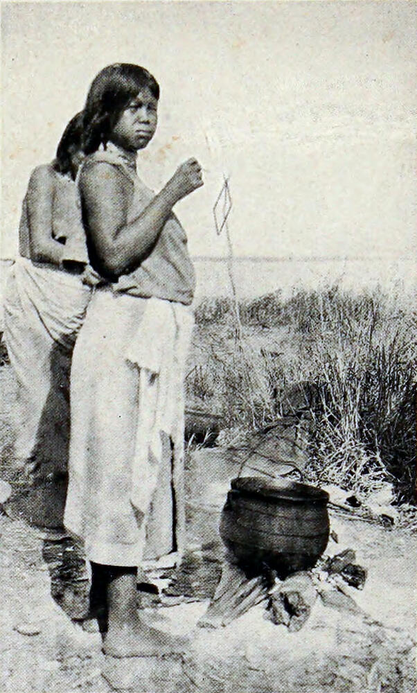 A photograph of a young girl standing beside a small black cooking pot on the ground.